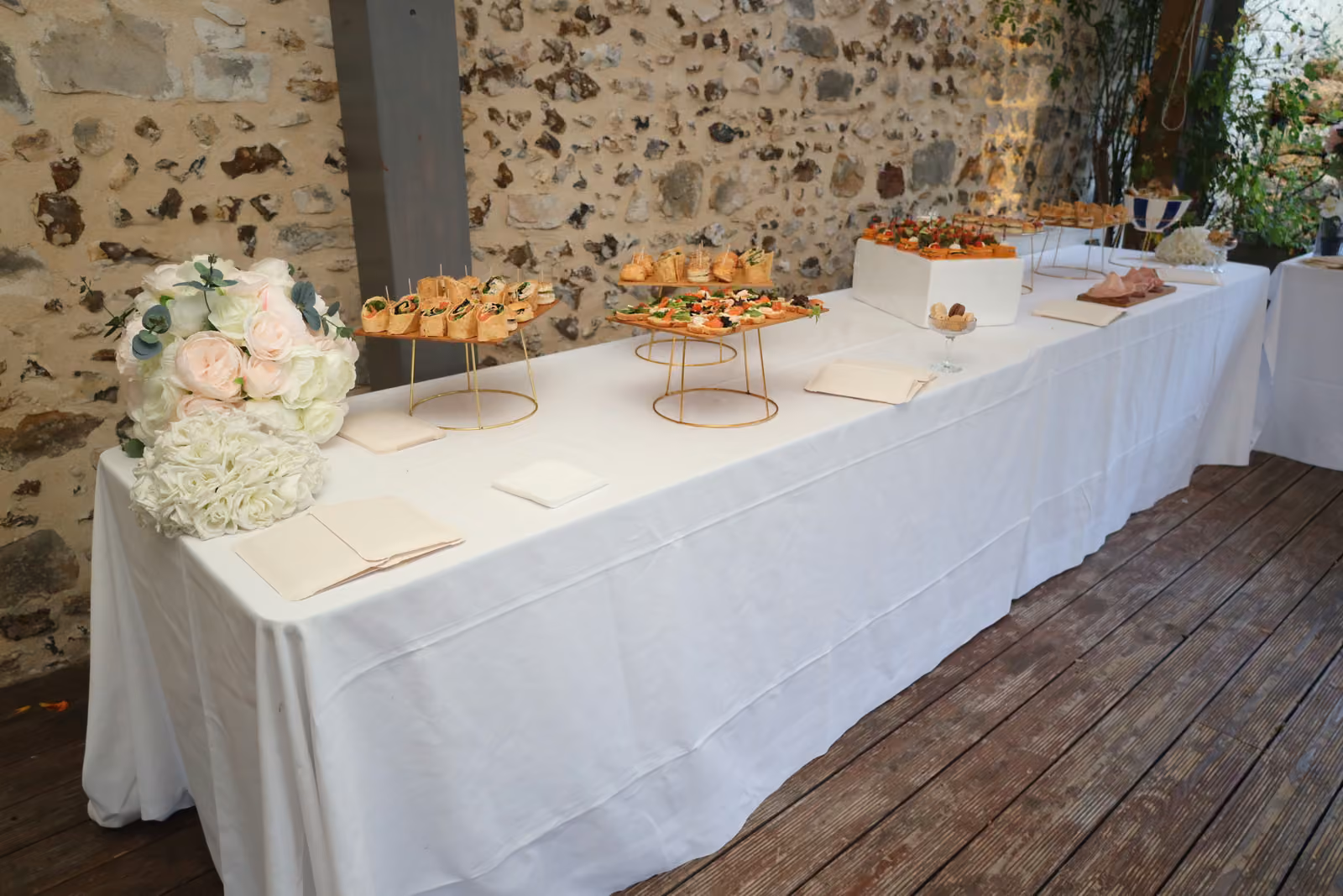 Buffet de cocktail en terrasse Buffet de cocktail sur nappe blanche en terrasse, présentoirs dorés garnis de bouchées et bouquet de roses