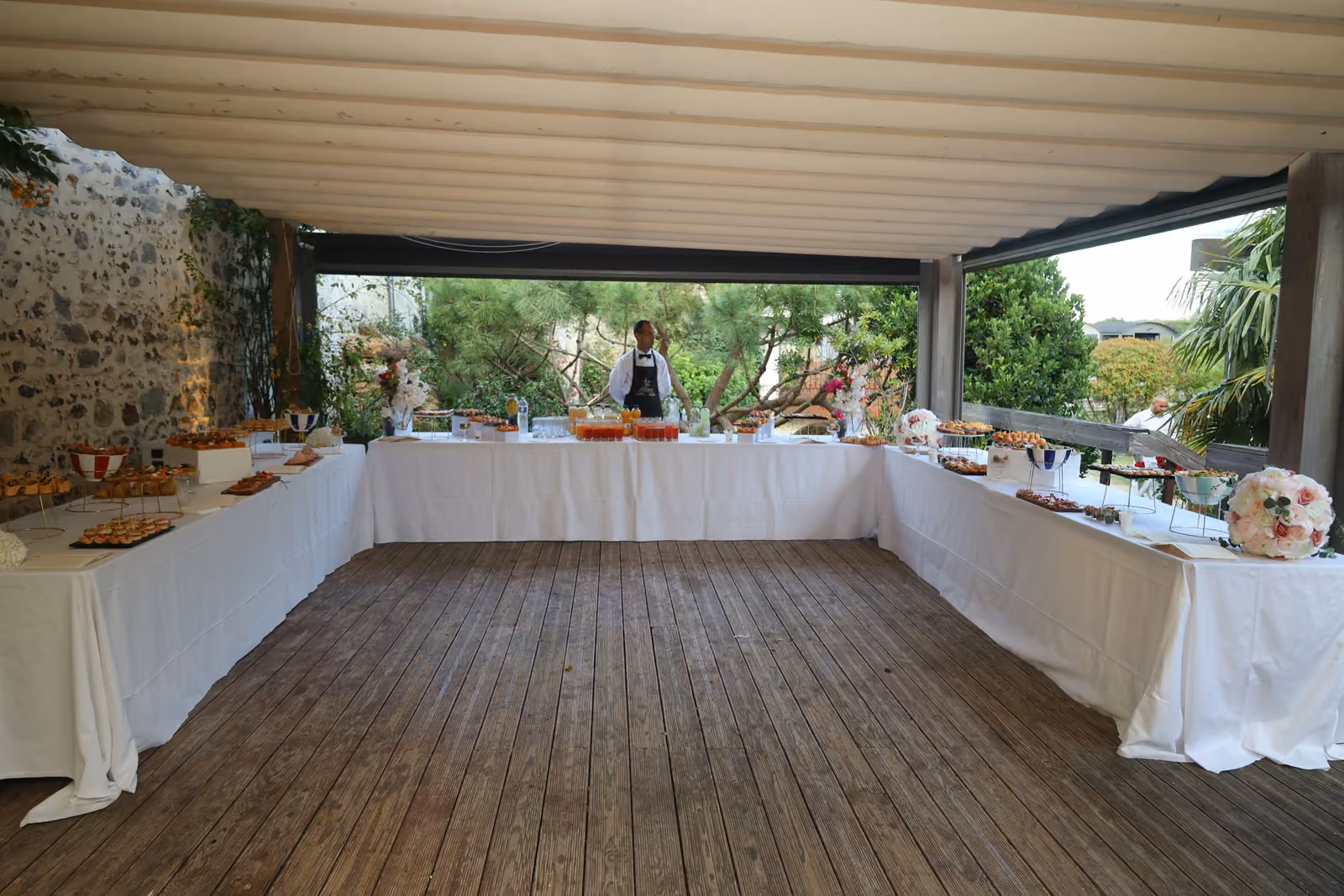 Buffet en U sous pergola Buffet en U sous une pergola, nappes blanches et parquet en bois, jardin en arrière-plan
