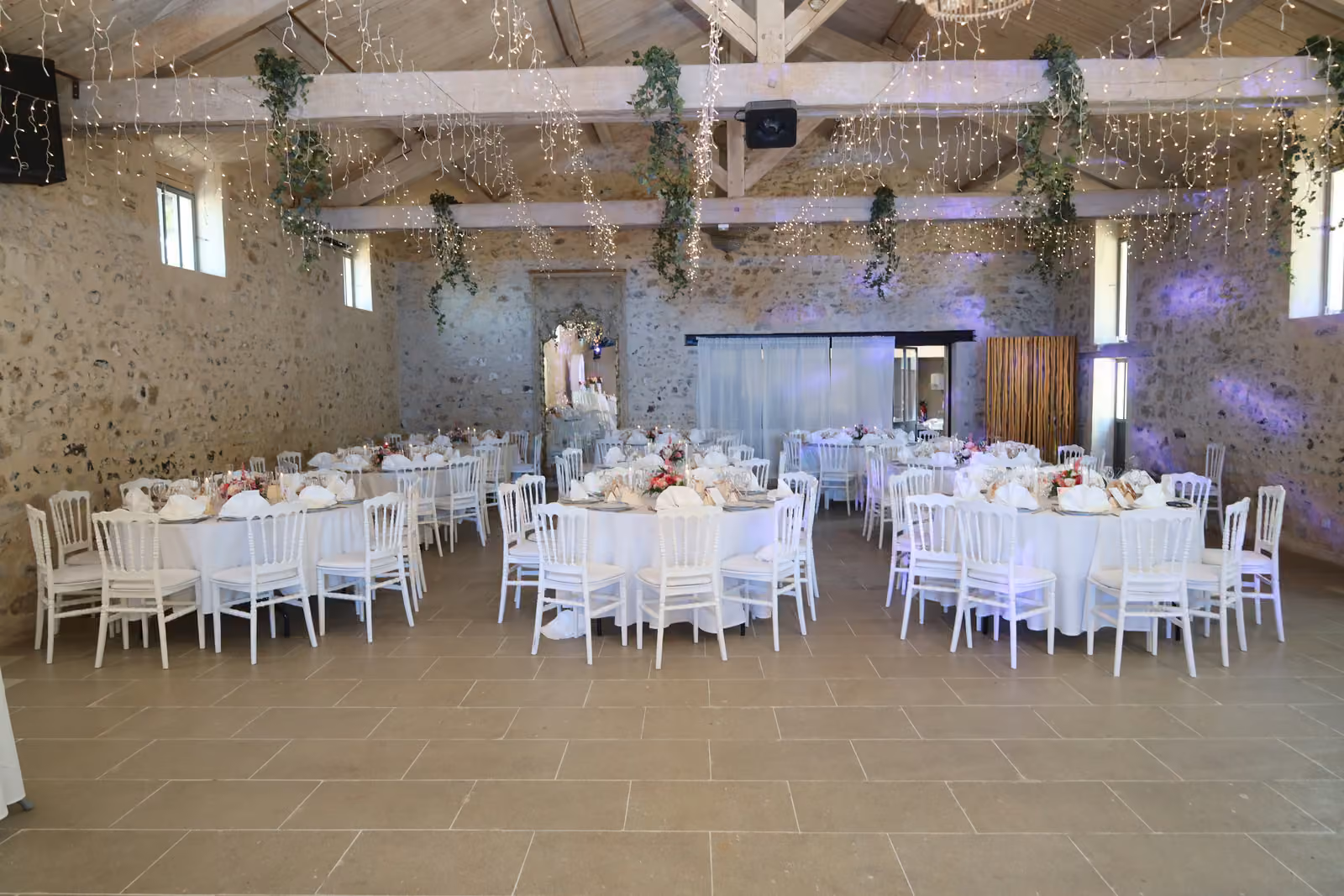 Salle de mariage dressée en tables rondes Salle de mariage avec tables rondes nappées de blanc et chaises Napoléon blanches sous une guirlande lumineuse
